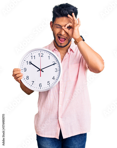 Handsome latin american young man holding big clock smiling happy doing ok sign with hand on eye looking through fingers