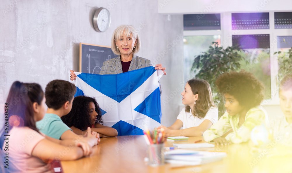 Fototapeta premium Friendly high school woman teacher tells pupils the history of Scotland in class and holds the national flag of the country .in her hands