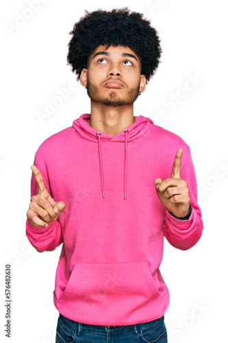 Young african american man with afro hair wearing casual pink sweatshirt pointing up looking sad and upset, indicating direction with fingers, unhappy and depressed.