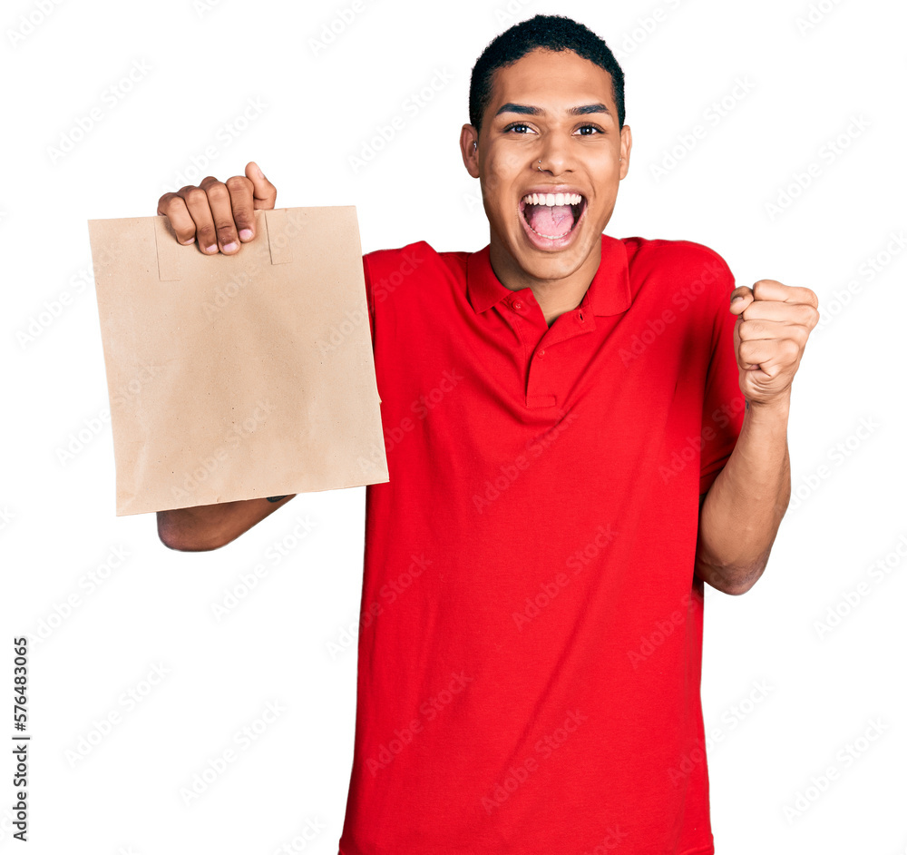 Young hispanic man holding take away paper bag screaming proud, celebrating victory and success very excited with raised arm Young hispanic man holding take away paper bag screaming proud, celebrating victory and success very excited with raised arm