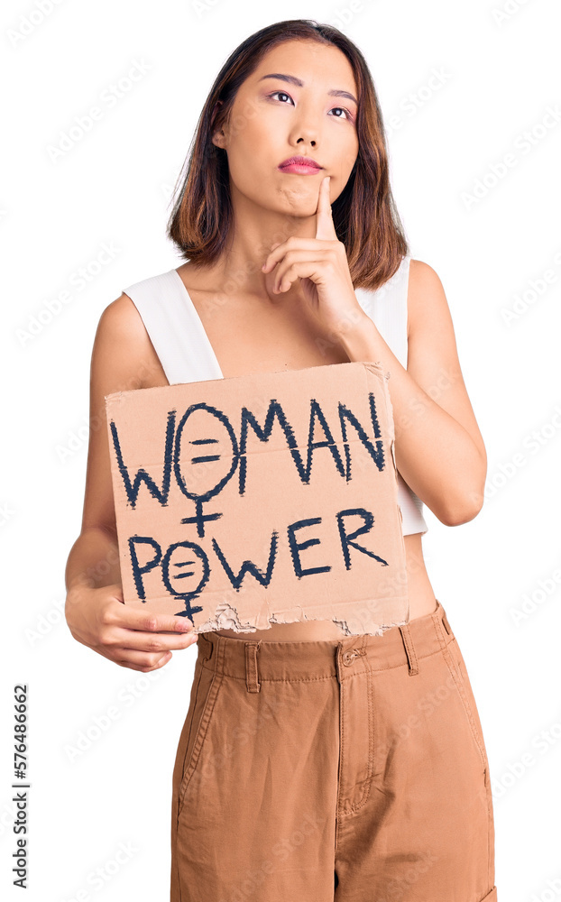 Young beautiful chinese girl holding woman power banner serious face thinking about question with hand on chin, thoughtful about confusing idea Young beautiful chinese girl holding woman power banner serious face thinking about question with hand on chin, thoughtful about confusing idea