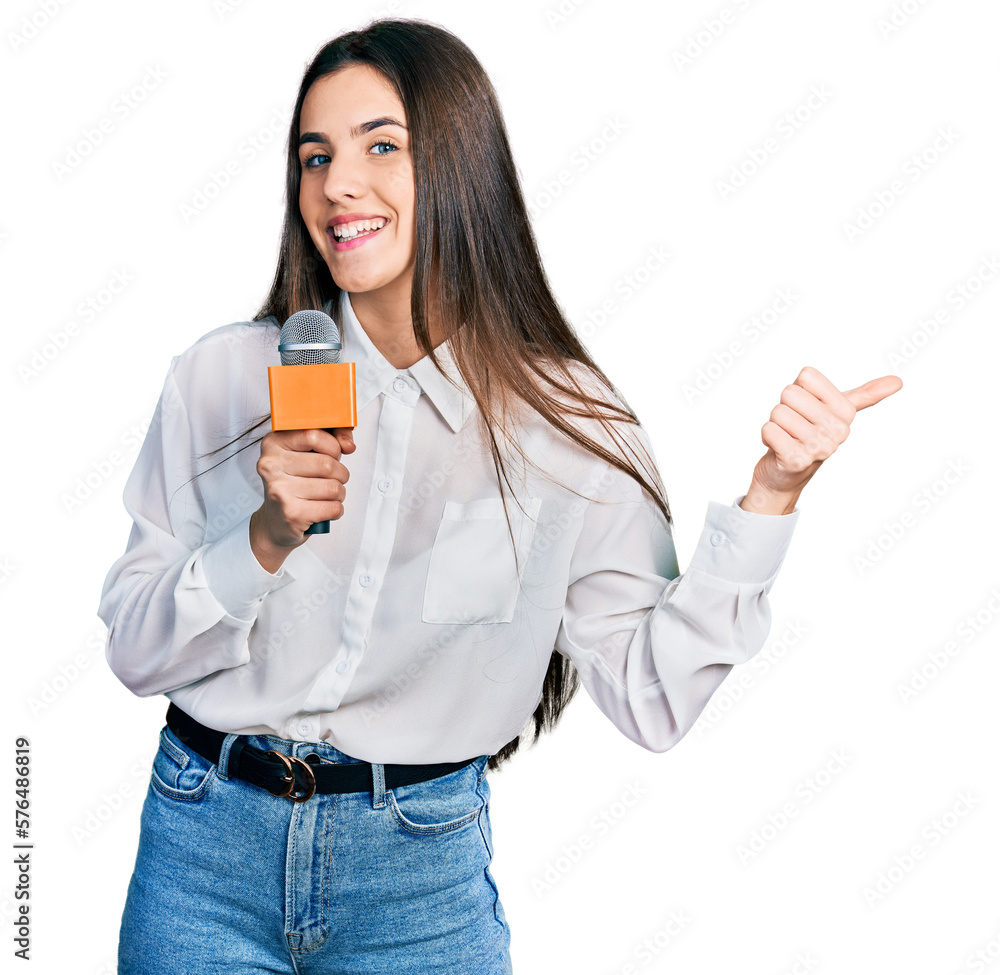Young brunette teenager holding reporter microphone pointing thumb up to the side smiling happy with open mouth Young brunette teenager holding reporter microphone pointing thumb up to the side smiling happy with open mouth