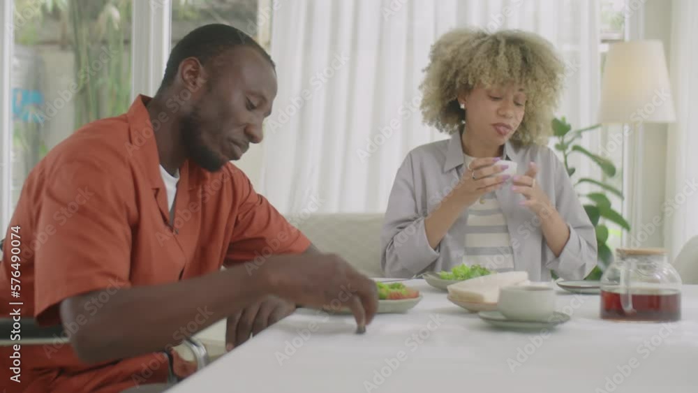 African American man with disability sitting in wheelchair, eating dinner and talking with wife while spending day together at home African American man with disability sitting in wheelchair, eating dinner and talking with wife while spending day together at home