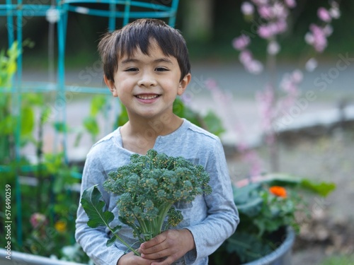 Adorable Asian American boy smiling and holding broccoli in garden 
