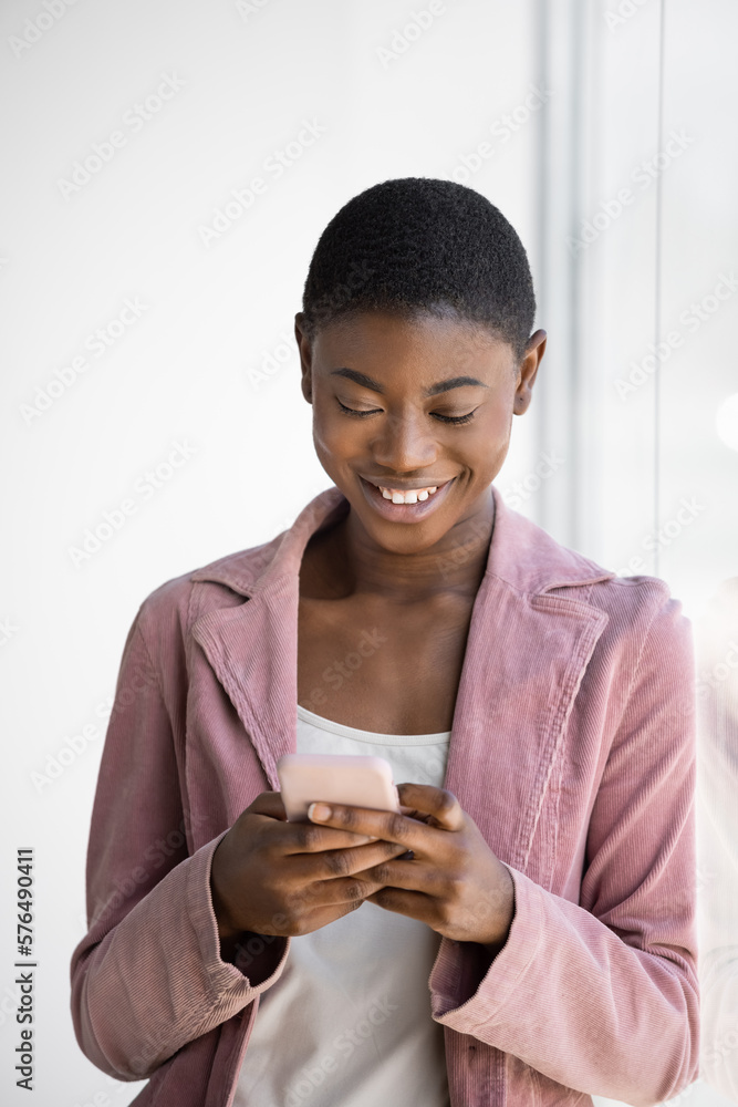 Smiling Black woman texting on the smartphone while leaning on glass window Smiling Black woman texting on the smartphone while leaning on glass window