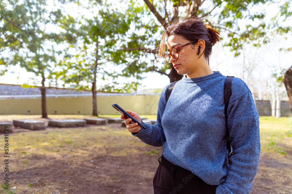 Obraz premium Joven mujer, con mochila, auriculares, usando el celular, telefono. Fotografía plano medio.