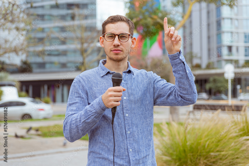 Fototapeta premium Hombre joven, caucásico, de 25 años, con lentes, con un micrófono y camisa azul al aire libre, posible reportero.