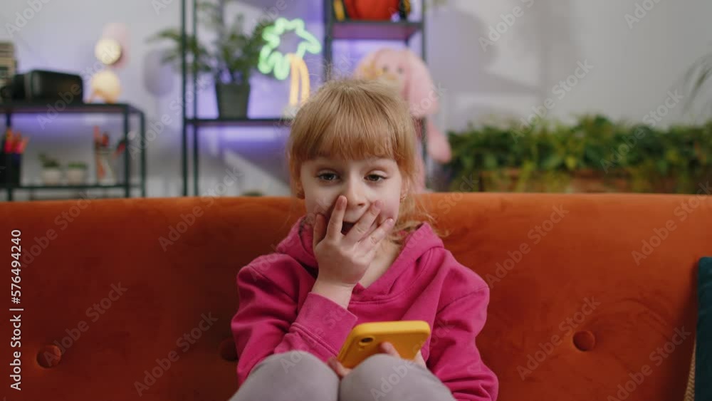Young blonde child kid girl use mobile smartphone typing playing browsing say Wow yes found out great big surprise, good news, celebrate online game win. Female toddler at home in room sits on sofa Young blonde child kid girl use mobile smartphone typing playing browsing say Wow yes found out great big surprise, good news, celebrate online game win. Female toddler at home in room sits on sofa