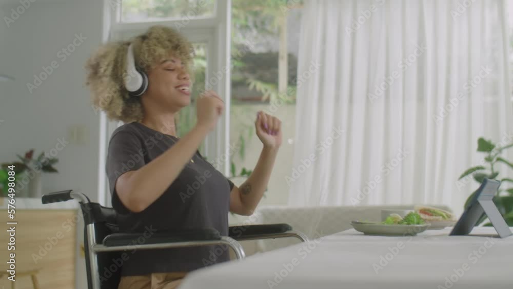 Young joyous woman with disability sitting in wheelchair, listening to music with headphones on tablet, smiling and dancing while having fun at home Young joyous woman with disability sitting in wheelchair, listening to music with headphones on tablet, smiling and dancing while having fun at home