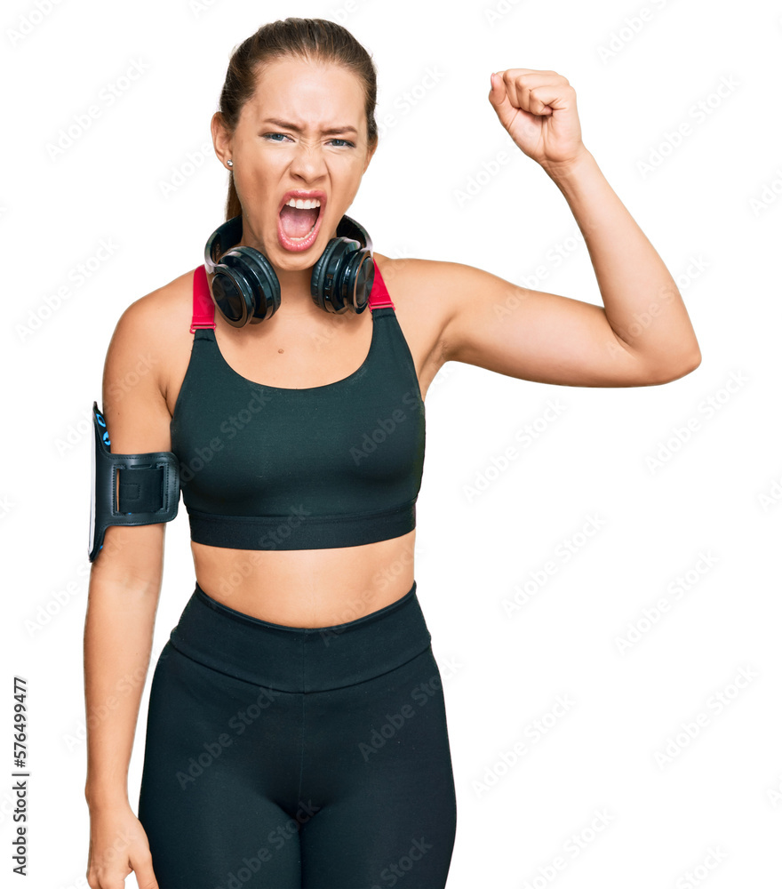Beautiful blonde woman wearing gym clothes and using headphones angry and mad raising fist frustrated and furious while shouting with anger. rage and aggressive concept.