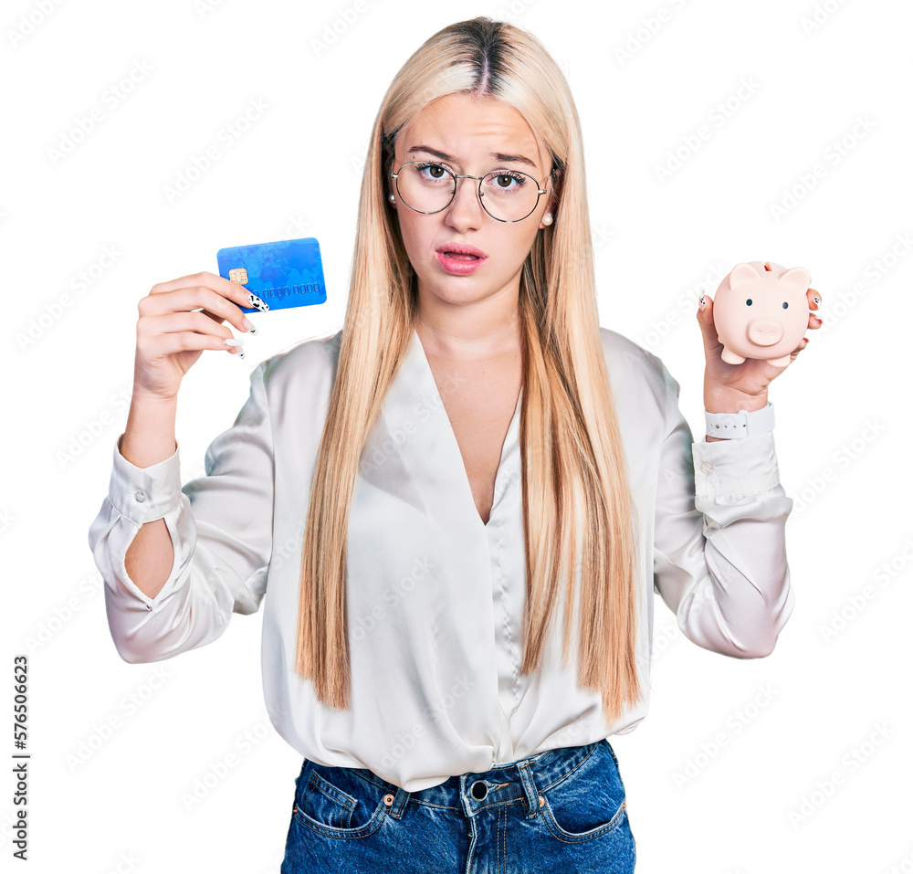 Beautiful blonde woman holding credit card and piggy bank in shock face, looking skeptical and sarcastic, surprised with open mouth Beautiful blonde woman holding credit card and piggy bank in shock face, looking skeptical and sarcastic, surprised with open mouth