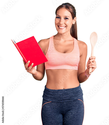 Young beautiful hispanic woman wearing gym clothes reading cooking recipe book holding spoon looking positive and happy standing and smiling with a confident smile showing teeth