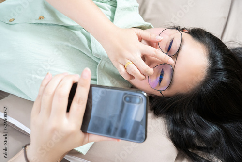 Beautiful glasses Asian woman lie on her back, chatting, typing, and texting, mobile in her bedroom with a pastel green - brown color theme. photo