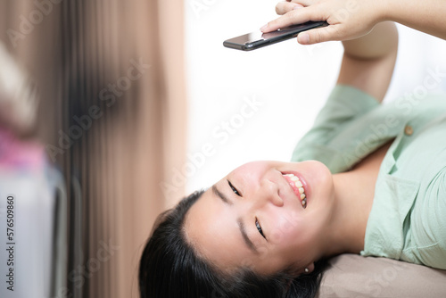 Beautiful Asian woman is lying down and playing mobile on the bed in her bedroom with a pastel green - brown color theme.