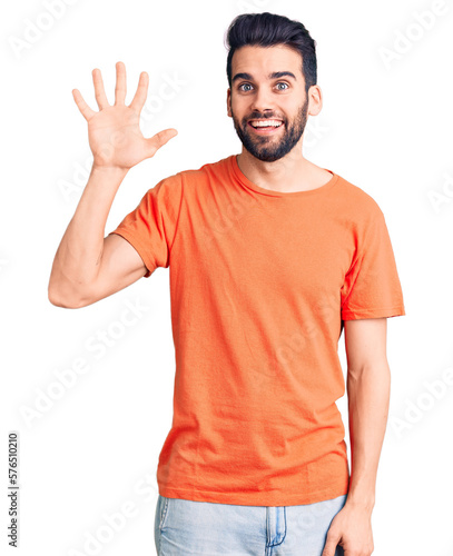 Young handsome man with beard wearing casual t-shirt showing and pointing up with fingers number five while smiling confident and happy.