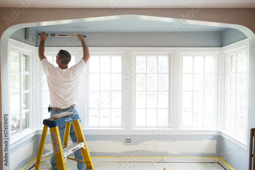 DYI Painter Carpenter Man Painting Overhead on Ladder Infront of Large Pain in Residential Family Living Space photo