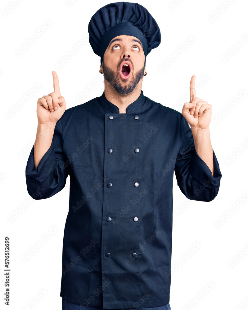 Young hispanic man wearing cooker uniform amazed and surprised looking up and pointing with fingers and raised arms.