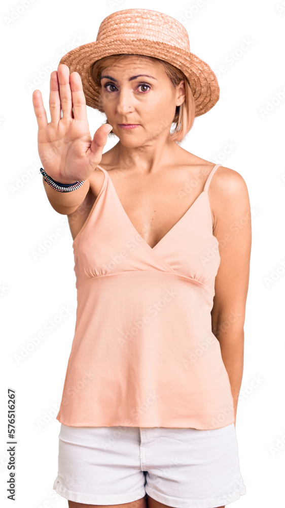 Young blonde woman wearing summer hat doing stop sing with palm of the hand. warning expression with negative and serious gesture on the face. Young blonde woman wearing summer hat doing stop sing with palm of the hand. warning expression with negative and serious gesture on the face.