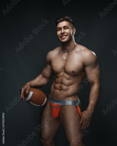 Cheerful handsome man with ball in studio. Naked smiling guy in orange jocks underwear with american football ball in his hand.