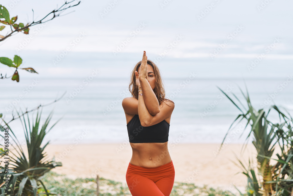 Eagle Pose Attractive athletic woman in black bra, red pants doing yoga exercises in front of sea Eagle Pose Attractive athletic woman in black bra, red pants doing yoga exercises in front of sea