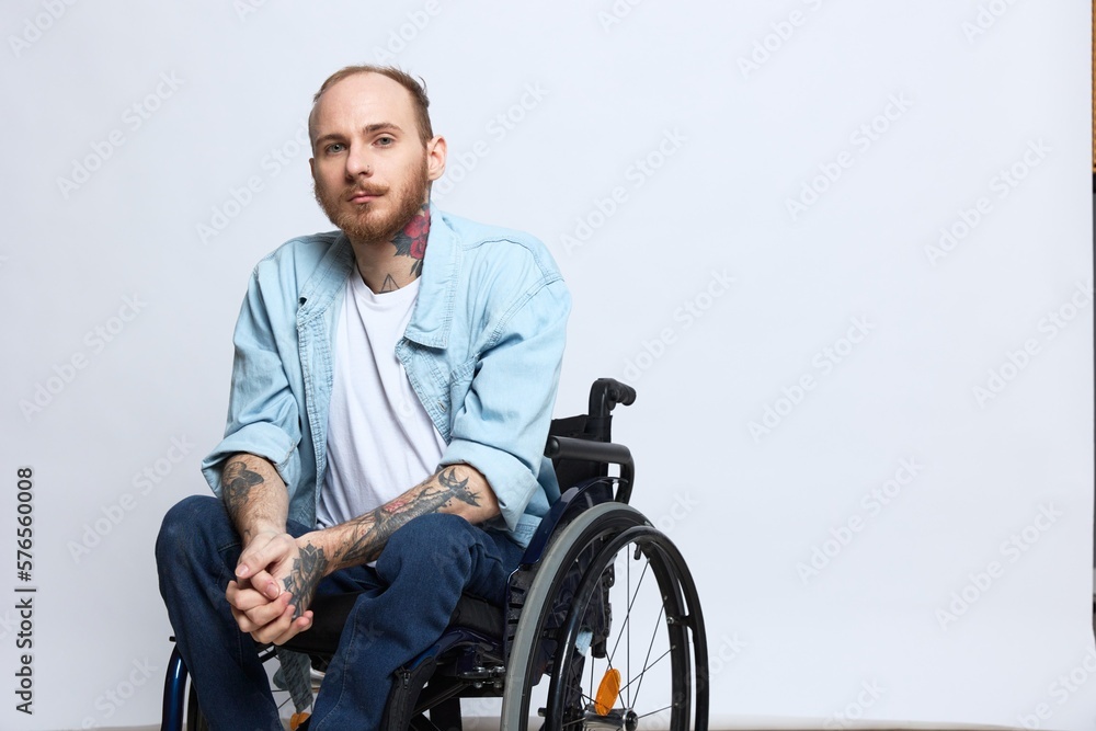 A man in a wheelchair looks thoughtfully at the camera, copy space, with tattoos on his arms sits on a gray studio background, the concept of health a person with disabilities, a real person A man in a wheelchair looks thoughtfully at the camera, copy space, with tattoos on his arms sits on a gray studio background, the concept of health a person with disabilities, a real person
