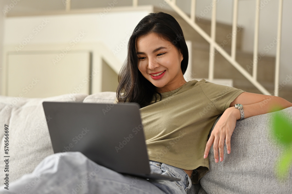 Happy young asian woman in casual clothes resting on couch and browsing internet on laptop. Happy young asian woman in casual clothes resting on couch and browsing internet on laptop.
