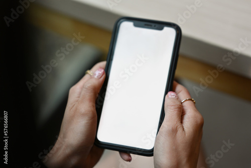 Close up view of woman hands holding smart phone while sitting at office desk. White screen for advertise design.