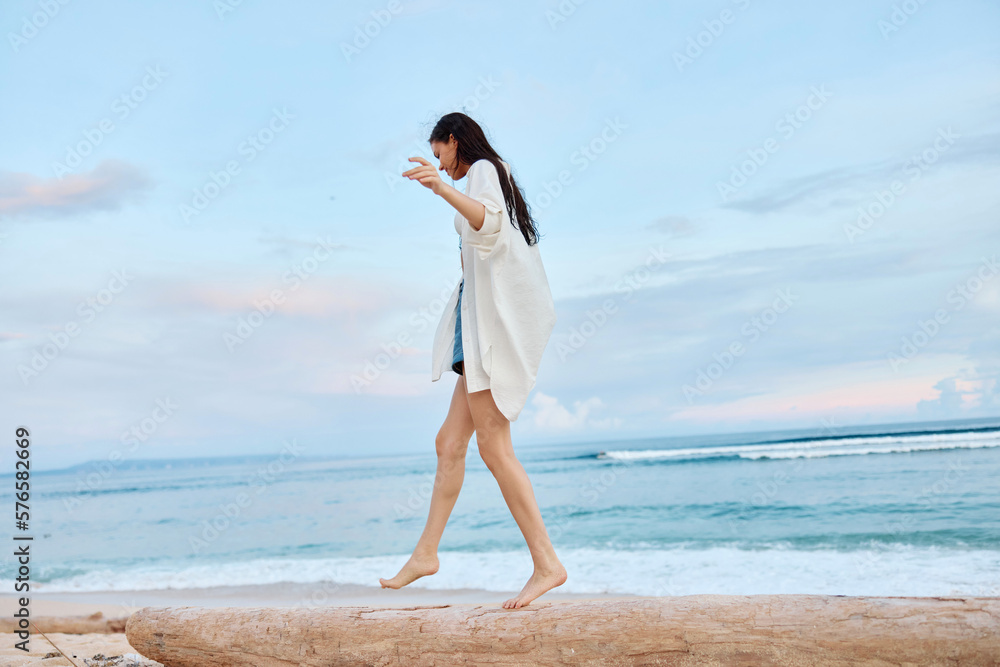Happy tanned woman in white swimsuit shirt and denim shorts walks the beach on a log on the sand by the ocean with wet hair after swimming, sunset light in Bali Happy tanned woman in white swimsuit shirt and denim shorts walks the beach on a log on the sand by the ocean with wet hair after swimming, sunset light in Bali