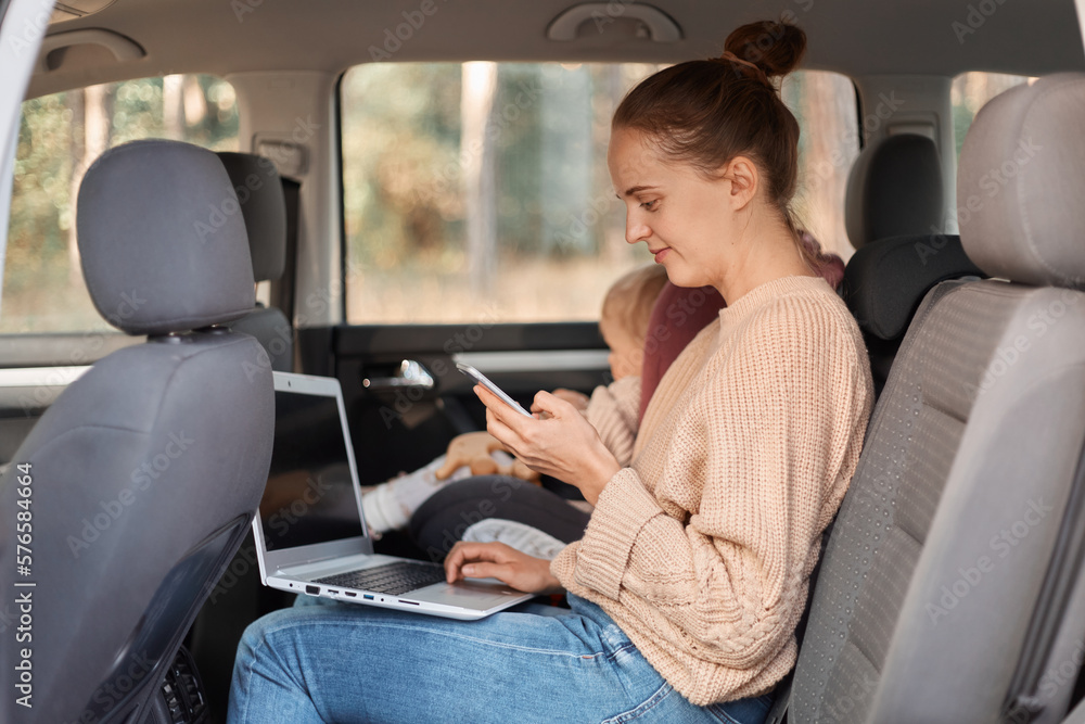 Image of cheerful young woman working on laptop while sitting with her baby daughter in safety chair on backseat of car. female freelancer using cell phone, smiling while looking at phone display.