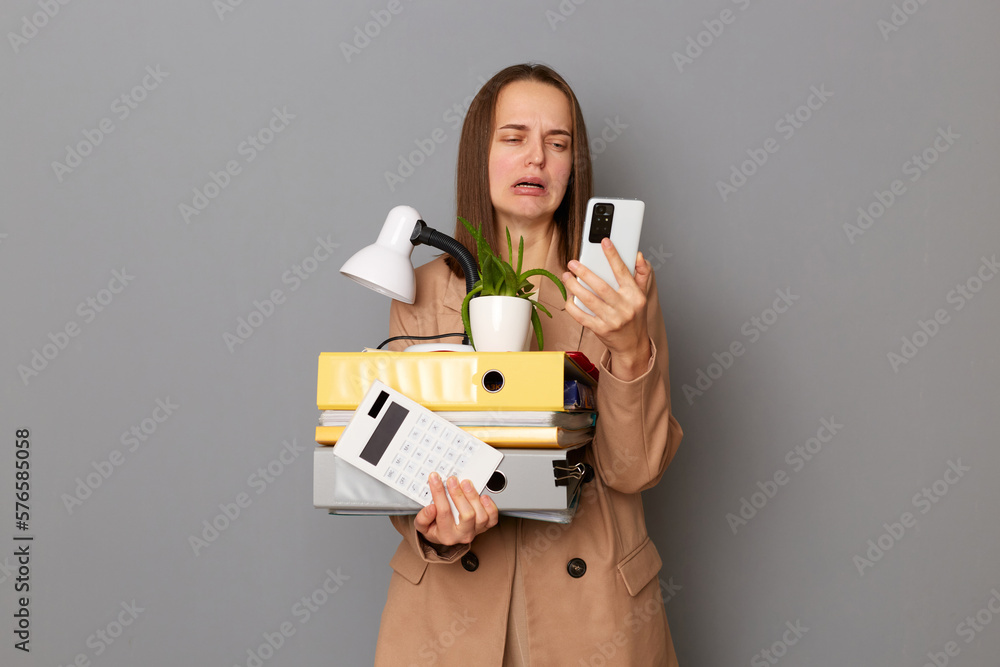 Indoor shot of sad fired businesswoman using mobile phone while standing isolated on gray background, holding belongings from office, looking at screen of phone and crying. Indoor shot of sad fired businesswoman using mobile phone while standing isolated on gray background, holding belongings from office, looking at screen of phone and crying.