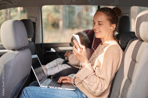 Profile portrait of woman wearing beinge jumper sitting with her baby daughter in safety chair on backseat of car, holding mobile phone in hands, listening music, having video call.