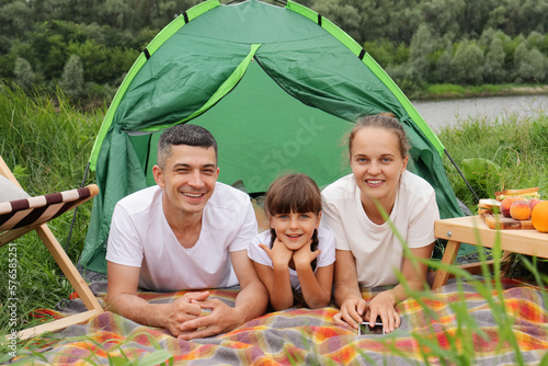 Outdoor shot of happy family, mother, father and cute little daughter with pigtails having picnic near the river, posing near green tent, looking smiling at camera.