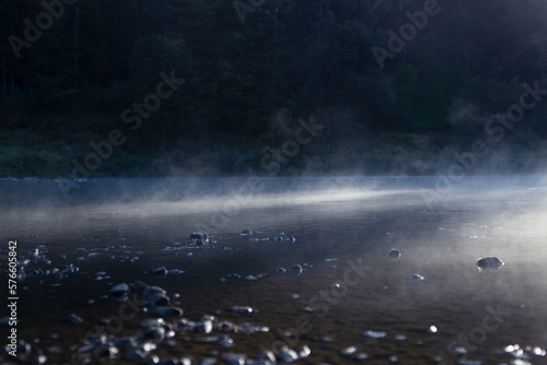 Stream of cold white fog above river water in sunbeams and deep blue silhouette of dark forest on shore  gloomy and sinister wild nature background.