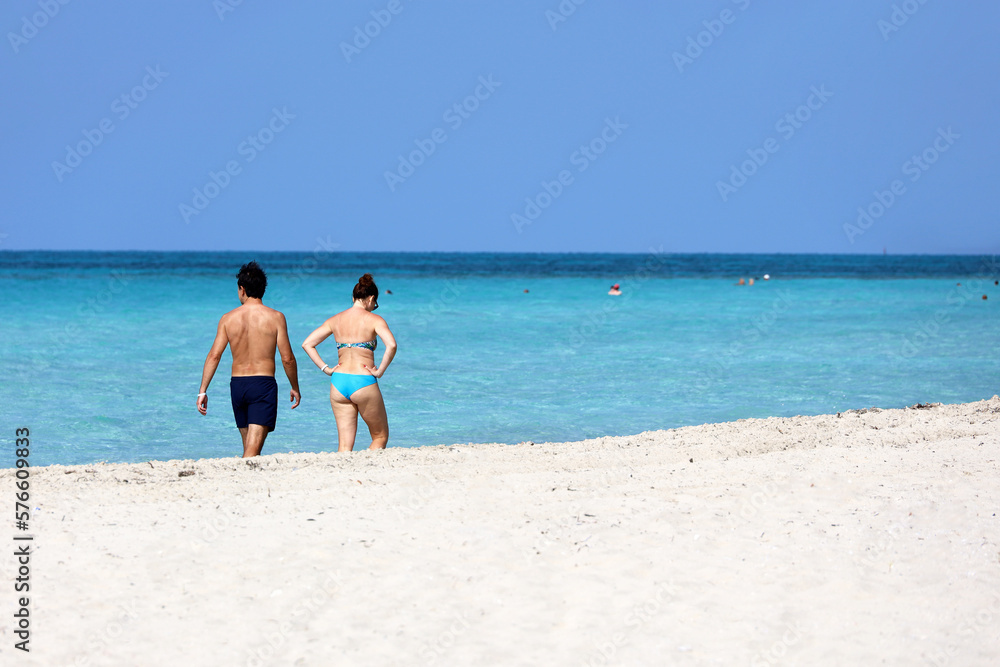 Couple in swimwear walking by white sand beach against the ocean waves. Man and woman together, romantic leisure on tropical coast