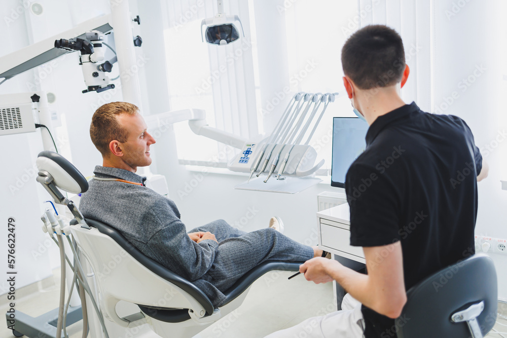 A young dentist doctor tells a male patient about dental care. A man was sitting in a dental chair at a doctor's appointment. Modern dental treatment A young dentist doctor tells a male patient about dental care. A man was sitting in a dental chair at a doctor's appointment. Modern dental treatment