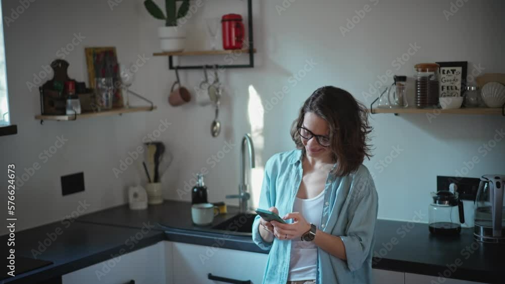 A young caucasian relaxed woman using her phone and drinking coffee while standing at the kitchen. A young caucasian relaxed woman using her phone and drinking coffee while standing at the kitchen.