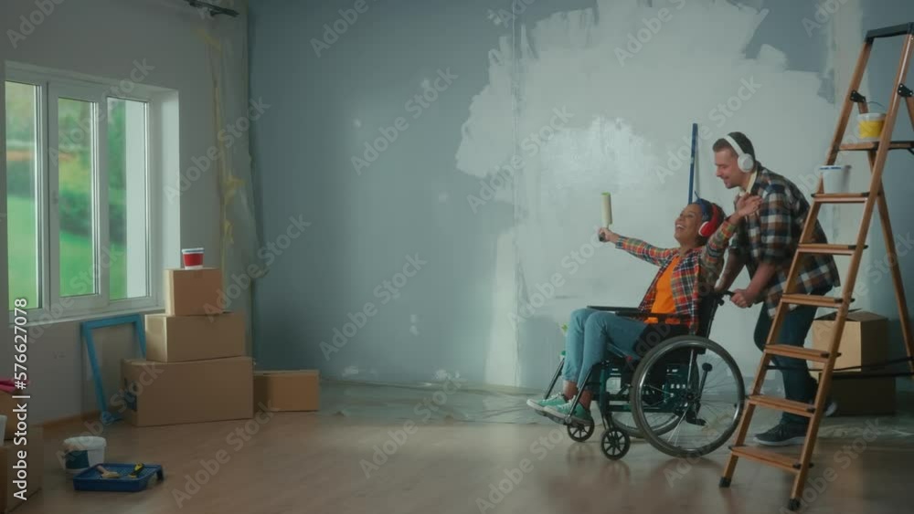 A white man wheeling an African American woman in a wheelchair into the room. A young couple in big headphones enjoys music, sings using a roller as a microphone and laughs. A white man wheeling an African American woman in a wheelchair into the room. A young couple in big headphones enjoys music, sings using a roller as a microphone and laughs.