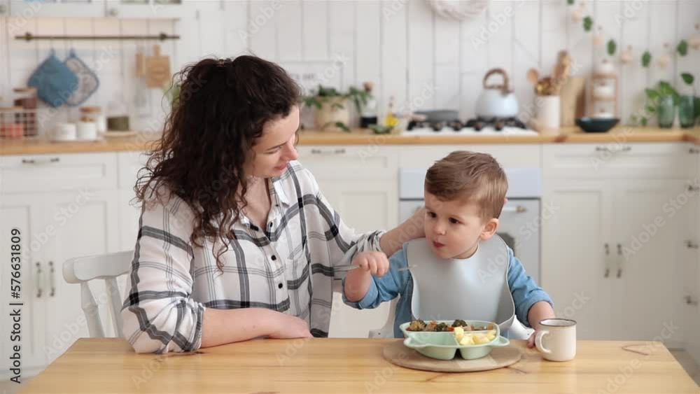 Young woman watches with delight as her son learns to eat on his own with a fork. Beautiful caucasian woman practices self-feeding with her baby. BLW. Dinner in a light and cozy kitchen Young woman watches with delight as her son learns to eat on his own with a fork. Beautiful caucasian woman practices self-feeding with her baby. BLW. Dinner in a light and cozy kitchen