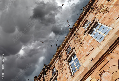 Dramatic storm clouds sky low angle view of stone facade decorated with diverse romantic bas relief and large crowd of pigeons flying on the blue sky background photo