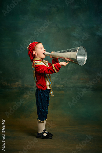 Little preschool age boy dressed up as medieval little prince and pageboy shouting at megaphone over dark vintage style background. Vintage fashion, emotions concept