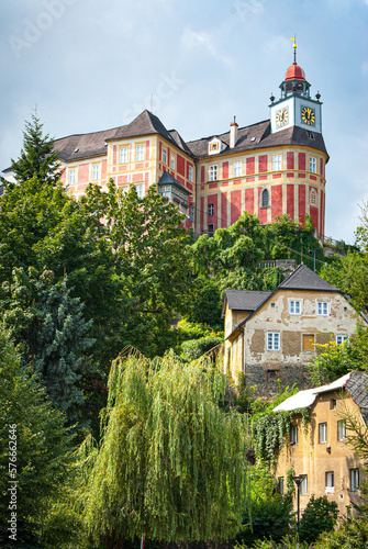 View of a pink chateau Jansky Vrch in Javornik, Czech Republic, sat on a steep hill, taken from the street level with trailing trees and crumbling houses in foreground

