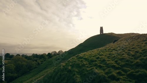 A drone shot of a hill in Somerset UK called Glastonbury Tor. The shot ascends to reveal fields that lead to the horizon. There is a tower in shot at the top of the hill. photo