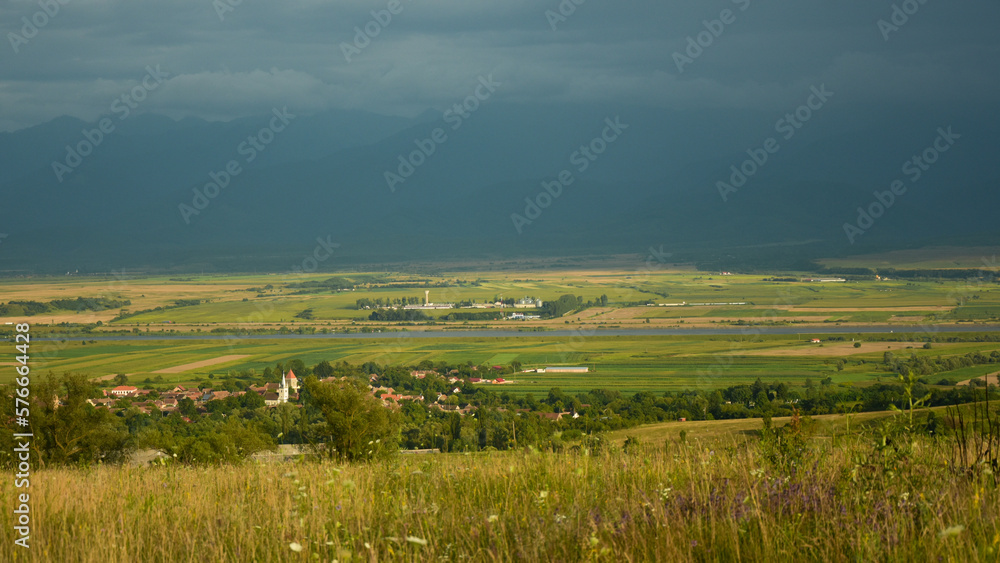 Scenic view of a Transylvanian village at twilight. The small town is located in a hilly area, surrounded by green fields, and a river. Storm is coming, black clouds gather in the sky. 