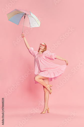 Full-length portrait of happy beautiful young girl in pink dress, smiling, posing with colorful umbrella over pink studio background. Concept of beauty, emotions, fashion, lifestyle and youth culture.