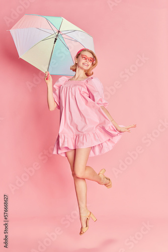 Summer vacation. Tender beautiful young girl in pink dress, sunglasses and colorful umbrella posing over pink studio background. Concept of beauty, emotions, fashion, lifestyle and youth culture.