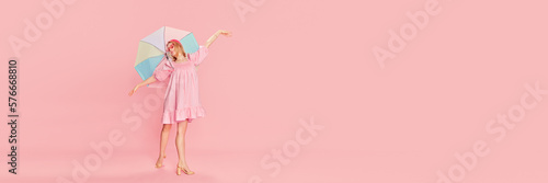 Happy beautiful young girl in pink dress, sunglasses and colorful umbrella posing over pink studio background. Concept of beauty, emotions, fashion, lifestyle, youth culture. Banner. Copy space for ad
