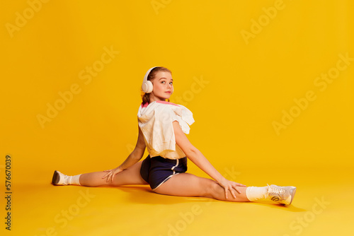 Hobby. Beautiful young girl in headphones and sports wear sitting on twine, training, stretching over yellow studio background. Concept of beauty, sportive lifestyle, hobby, emotions. Ad © master1305