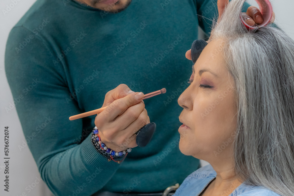 Fototapeta premium Stylist applying makeup in a professional makeup session on a senior adult woman, long gray hair, warm colors, lips with pink lipstick, beauty salon. Fashion and beauty concept