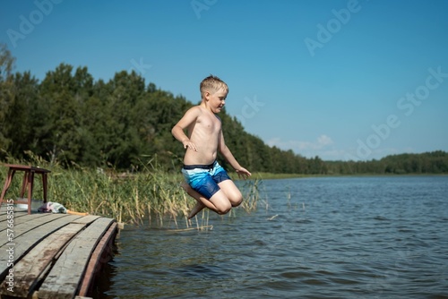 cute caucasian boy jumping from wooden pier diving into lake in countryside