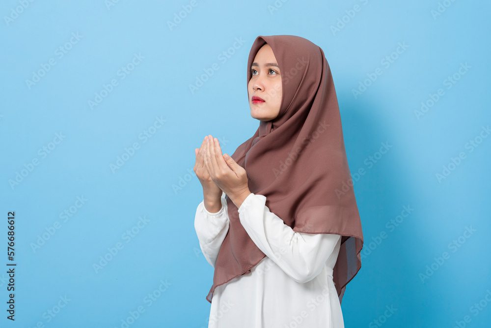 Happy muslim woman doing praying gesture looking up with a hopeful face isolated on blue background Happy muslim woman doing praying gesture looking up with a hopeful face isolated on blue background